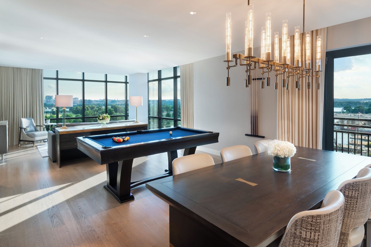 A modern hotel room with a pool table, a dining table with chairs, and large windows offering a city view.