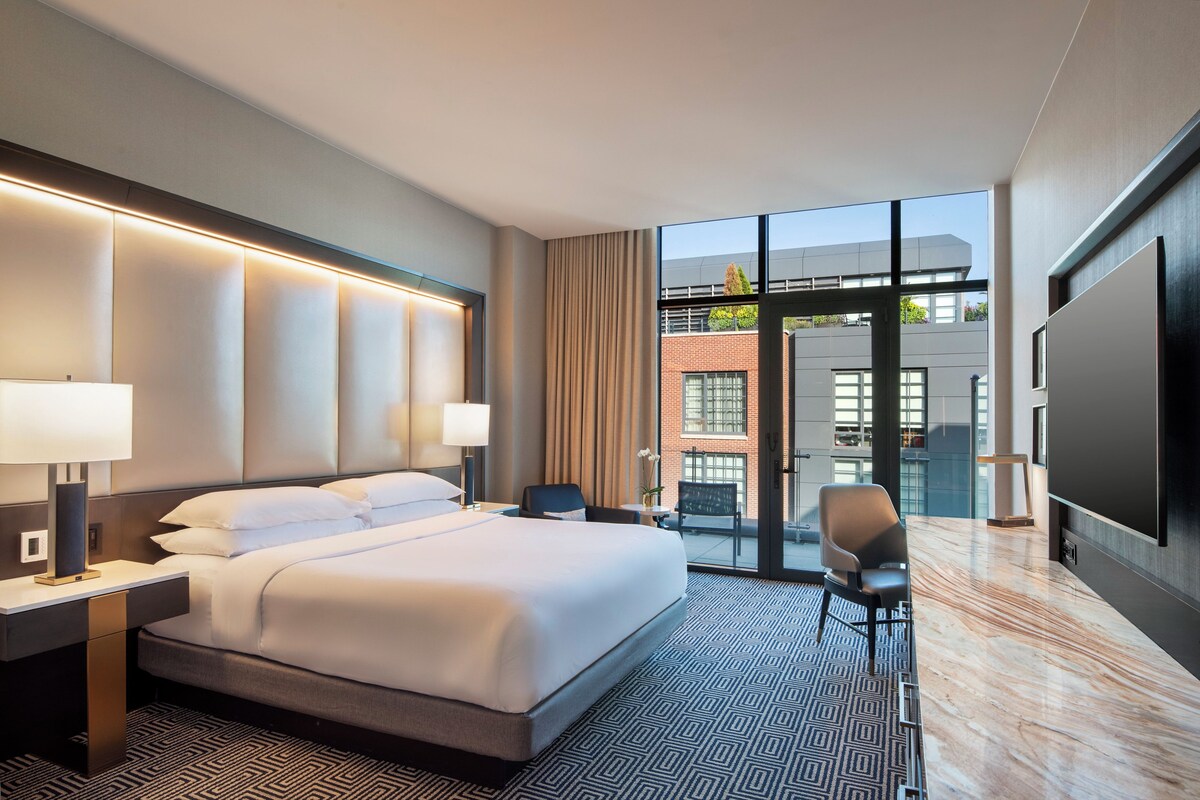 Una habitación de hotel moderna con una cama grande, dos mesitas de noche, un televisor de pantalla plana y vistas a la ciudad a través de amplias ventanas.
