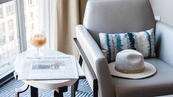 A hotel room with a grey armchair, a small table with a newspaper, a glass of drink, and a lamp.