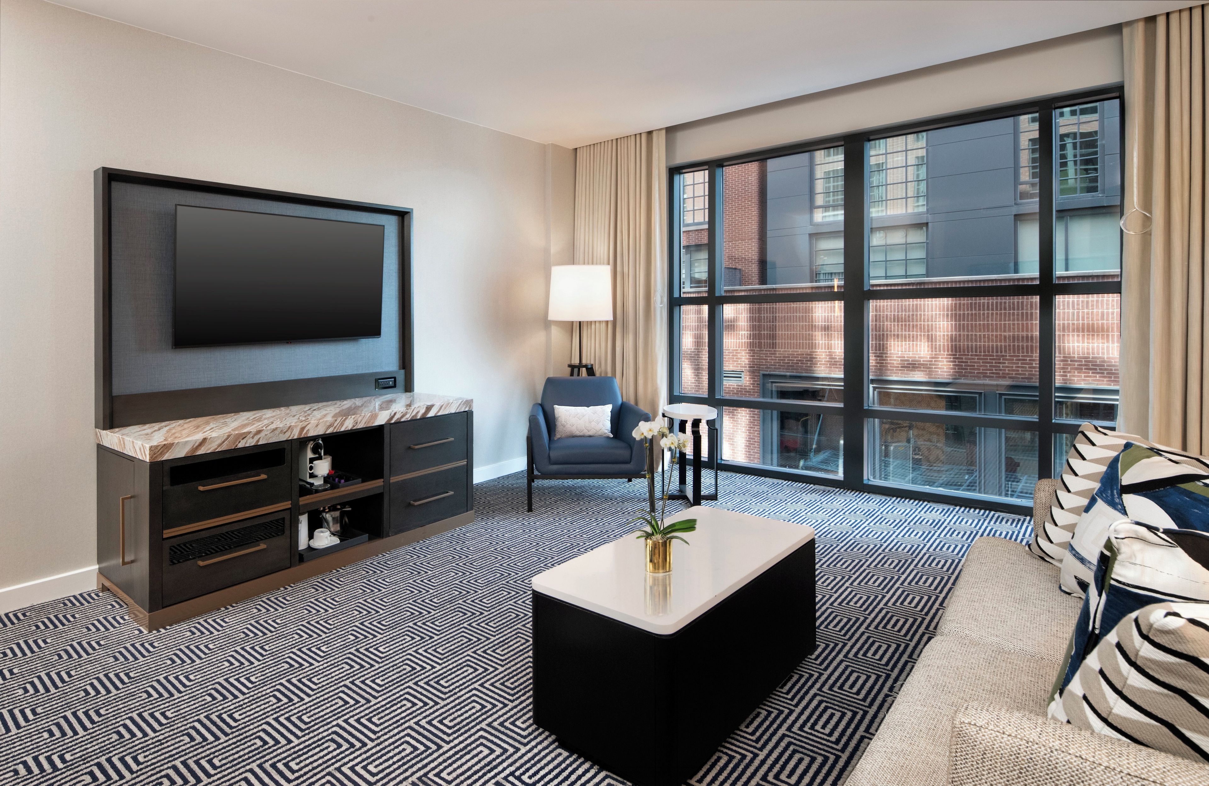 A modern hotel room with a large TV, a sofa, a coffee table, and a view of the city through large windows.