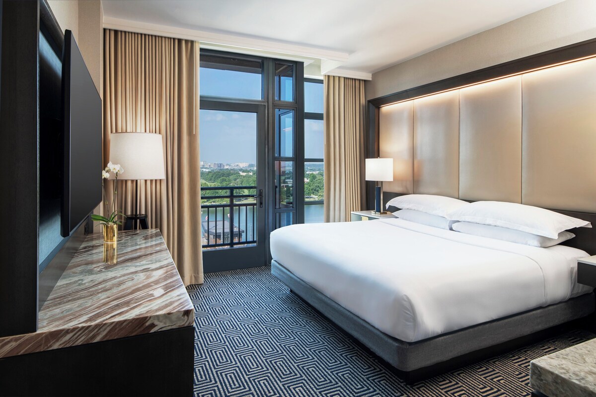 A hotel room with a bed, a desk with a lamp, a view of the city, and a large window.