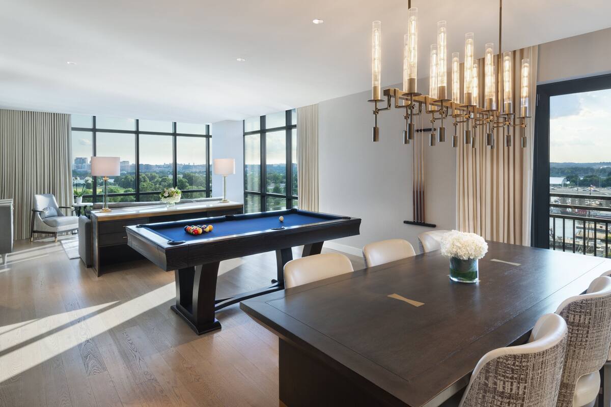 A modern hotel room with a pool table, a dining table with chairs, and large windows offering a city view.