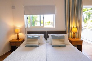 A double bed with two nightstands, each with a lamp, a window with blinds, and a view of greenery outside. - Centre Algarve (Olhao)