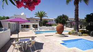 Zona de piscina con sillas de descanso y sombrillas, una maceta decorativa grande y un edificio de fachada blanca y tejado rojo.