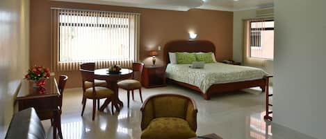 A hotel room with a bed, a sofa, a dining table with chairs, a small table with a laptop, and a window with blinds.