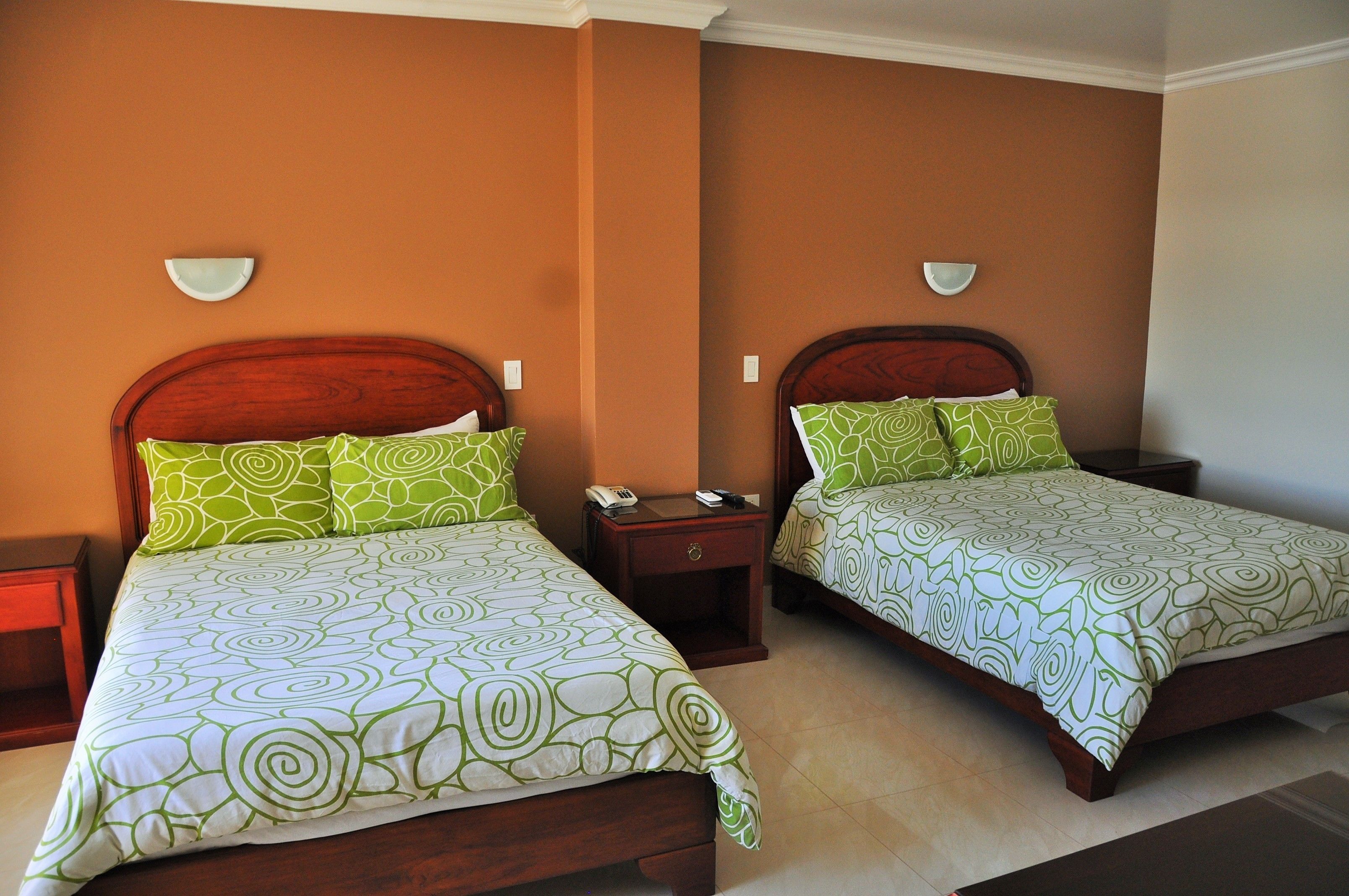 Dos camas matrimoniales con ropa de cama verde estampada, cabeceros de madera, mesitas de noche con lámparas y apliques de pared.