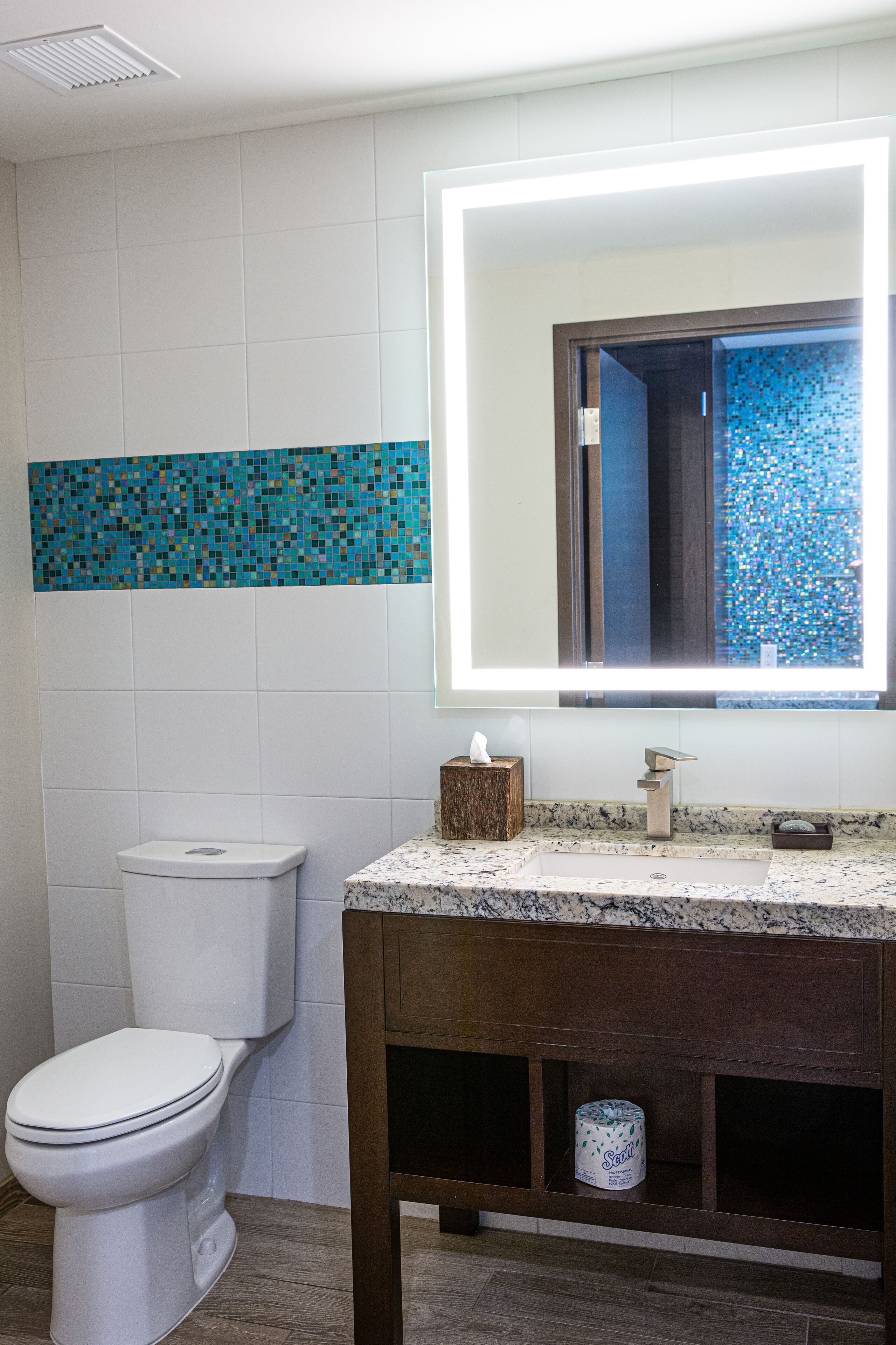 Un baño con inodoro, un lavamanos con encimera de mármol, tocador de madera y espejo con revestimiento de mosaico azul.