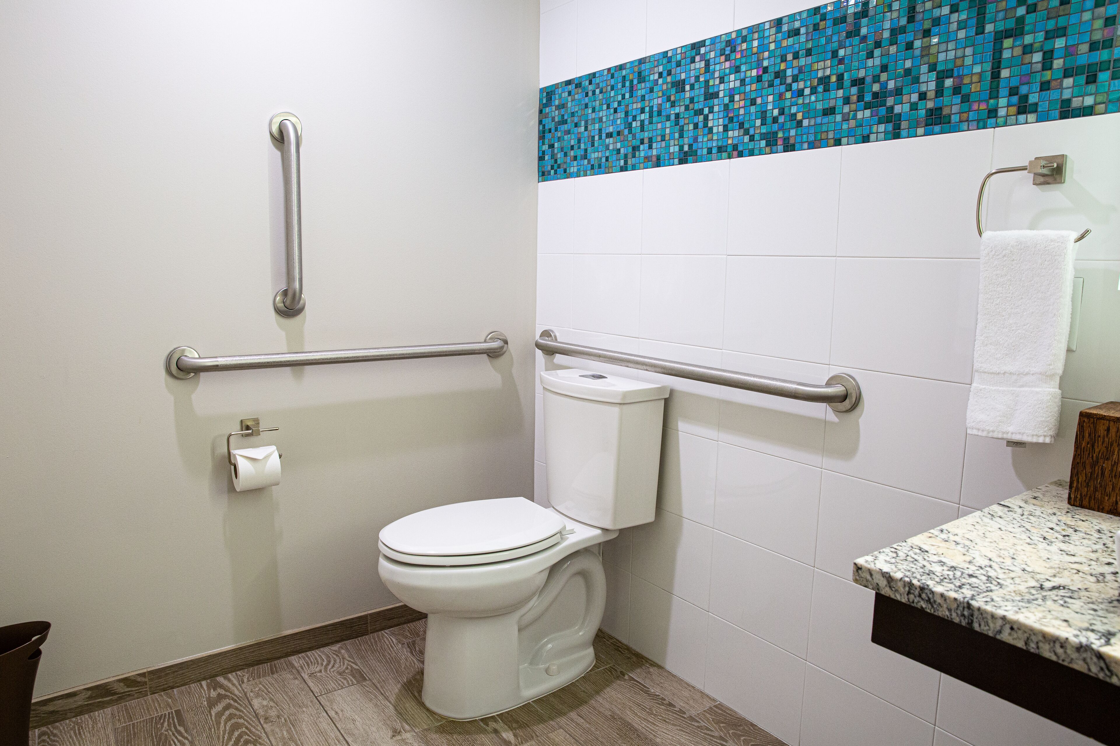 A bathroom with a white toilet, grab bars, a blue mosaic tile backsplash, and a marble countertop.
