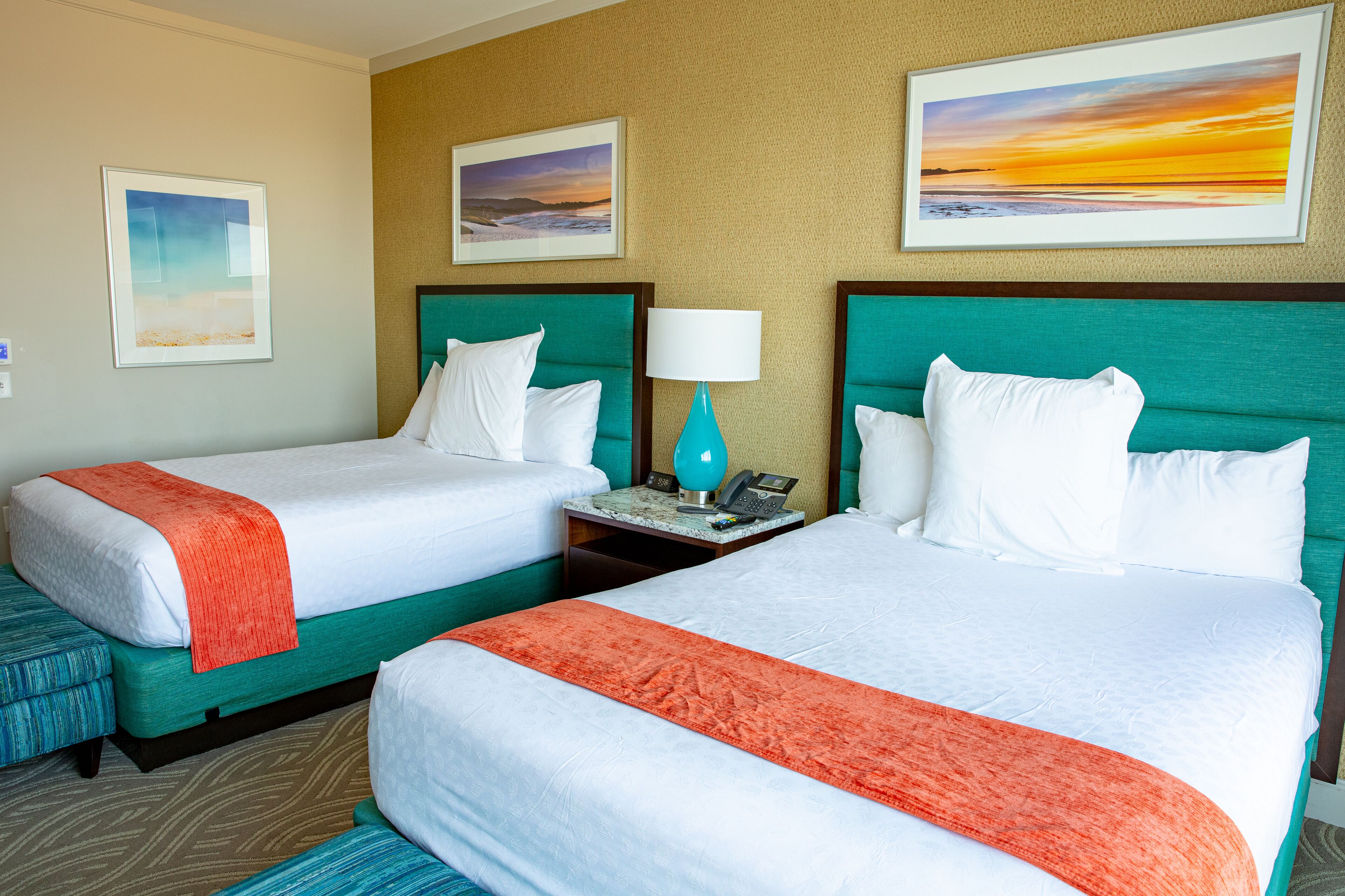 A hotel room with two beds, a nightstand with a lamp, and two framed pictures on the wall.