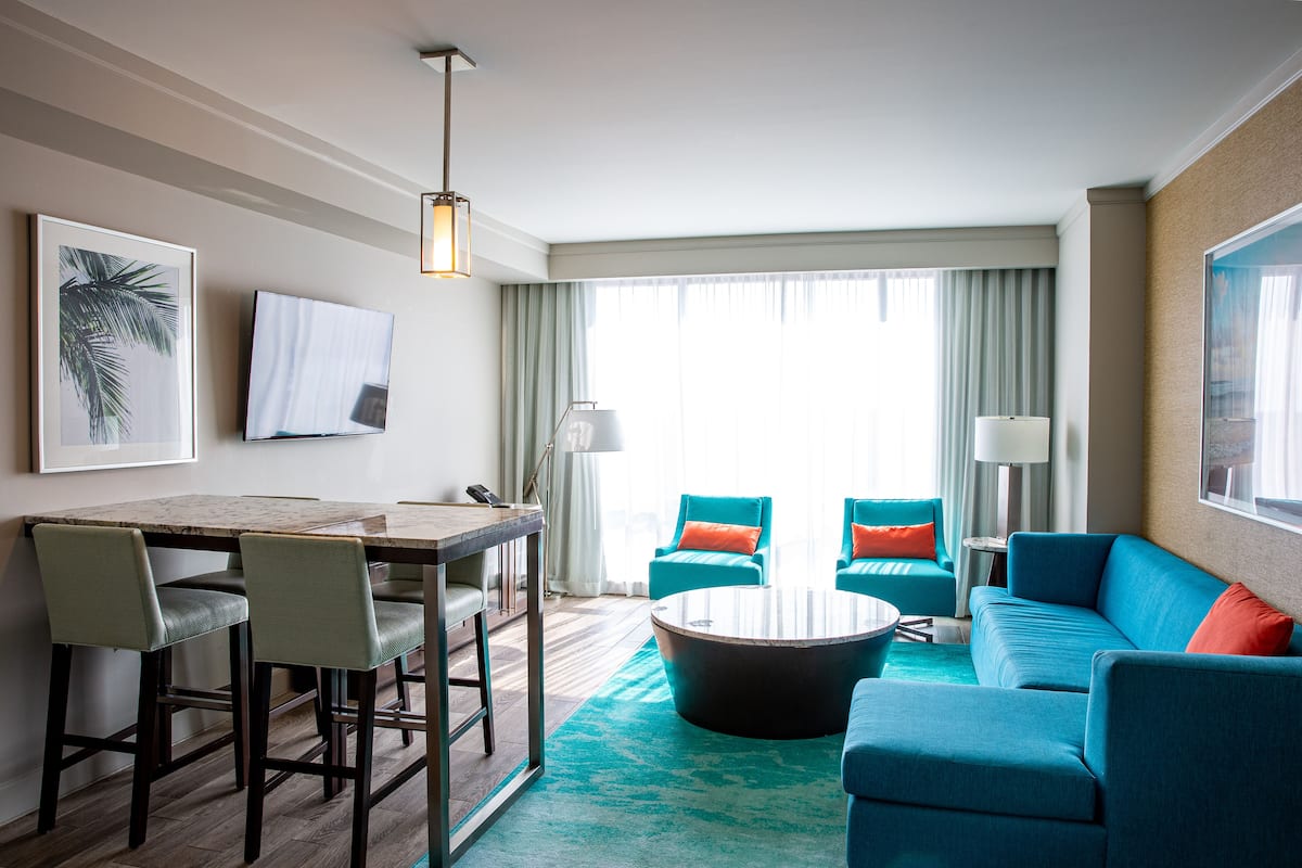 Ein modernes Hotelzimmer mit Essbereich, einer Sofagarnitur und einem großen Fenster mit Vorhängen.