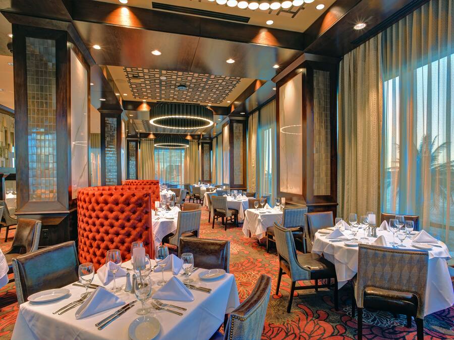 A hotel dining area with multiple tables set for dining, a large orange cushioned chair, and a patterned carpet.