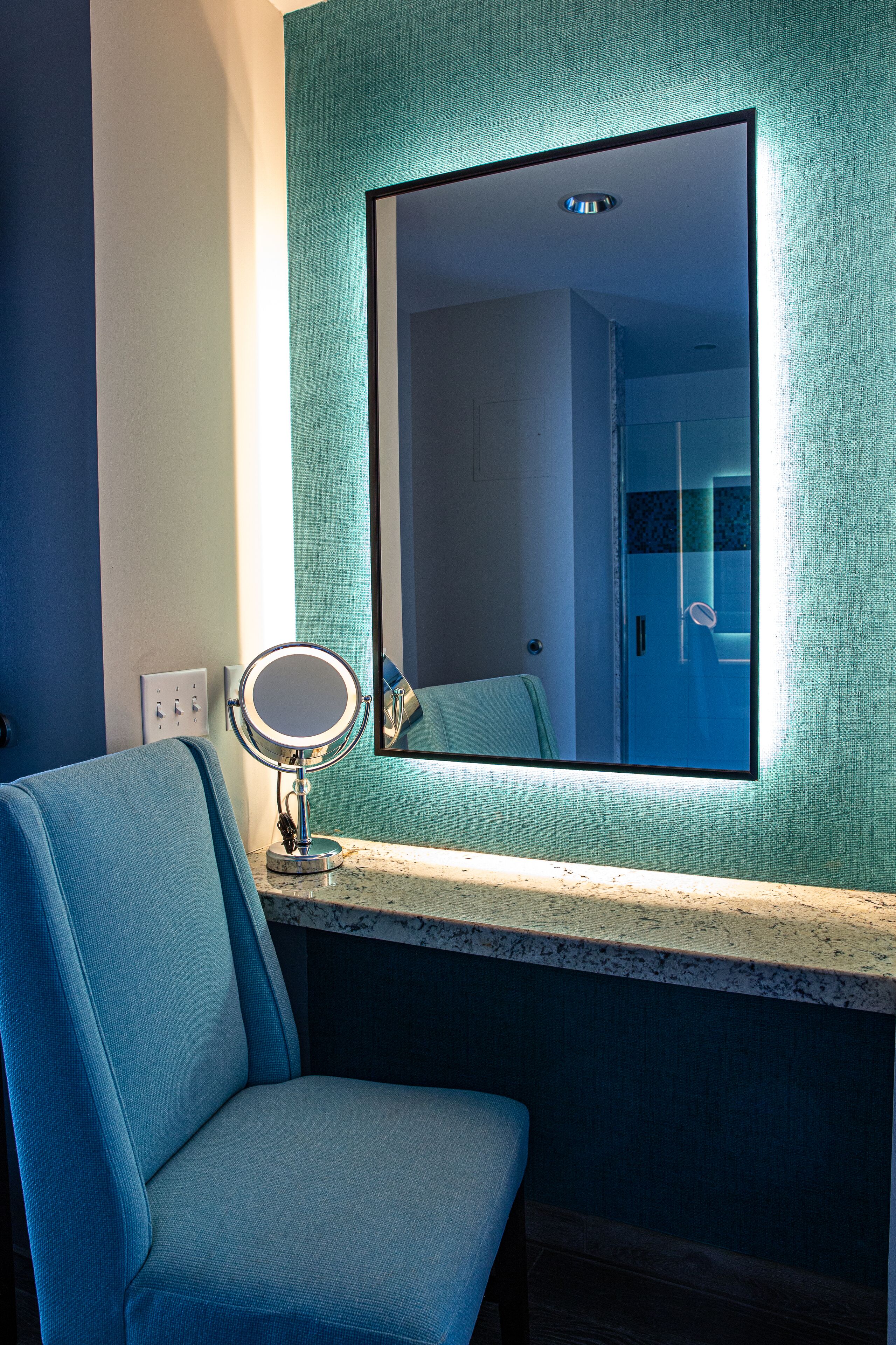A bathroom with a blue chair, a round mirror, and a large mirror on the wall.