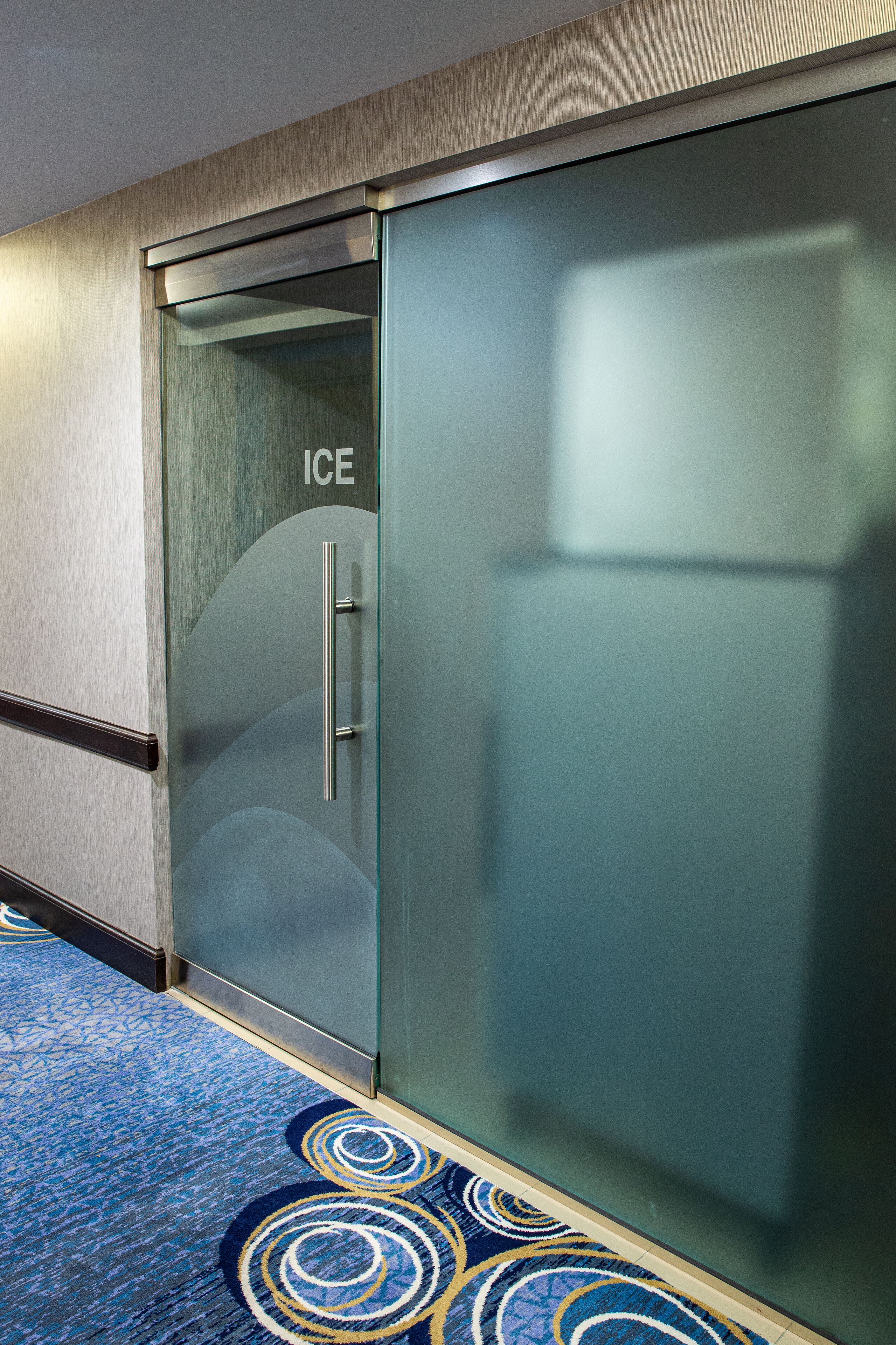 A hotel room with a glass door labeled "ICE," a blue patterned carpet, and a wooden floor.