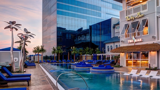 Ein Hotelpoolbereich mit Liegestühlen, einer Tiki-Bar und einem modernen Gebäude im Hintergrund.