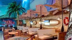 Ein Restaurant mit Holztischen und -sitzen, einem Wandbild mit tropischer Szenerie und hängenden Holzdekorationen.