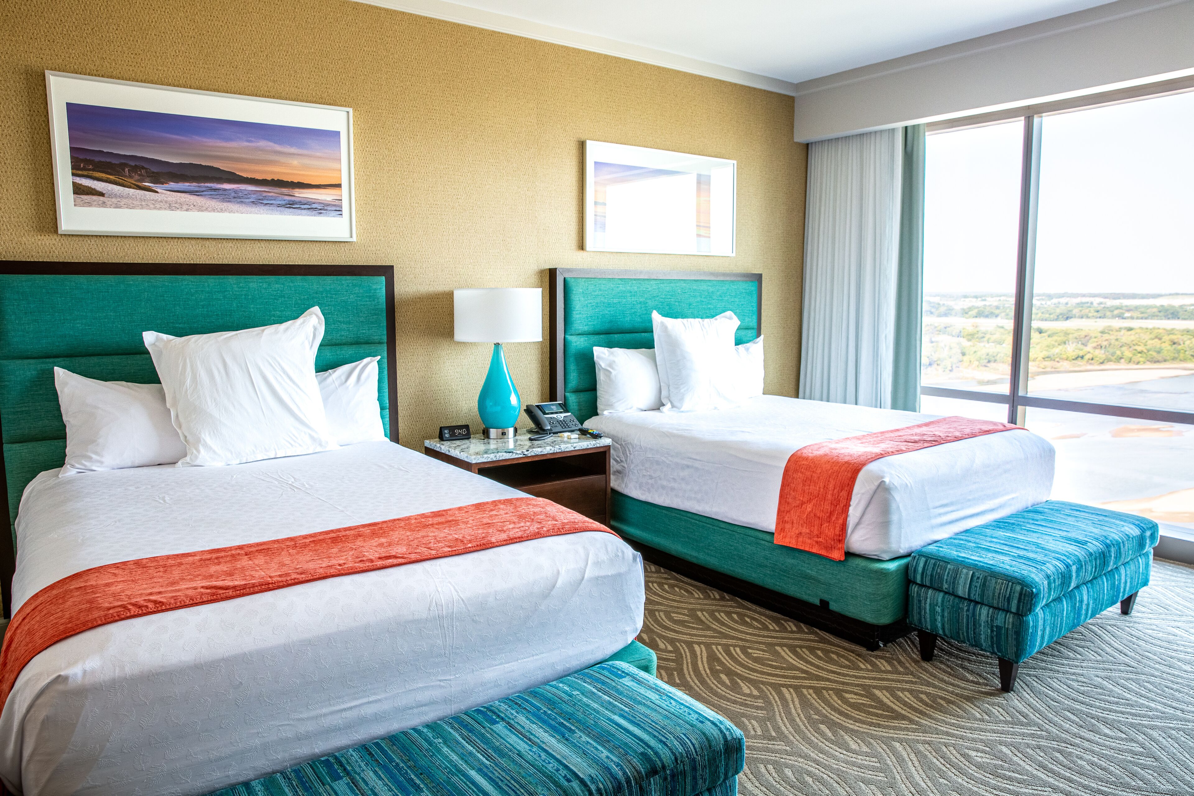 A hotel room with two beds, a nightstand, a lamp, and a large window with a view.