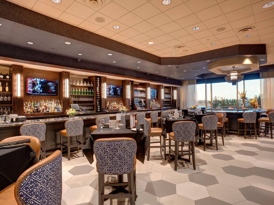 A bar area with multiple seating options, a television screen, and a variety of bottles and glasses on shelves.