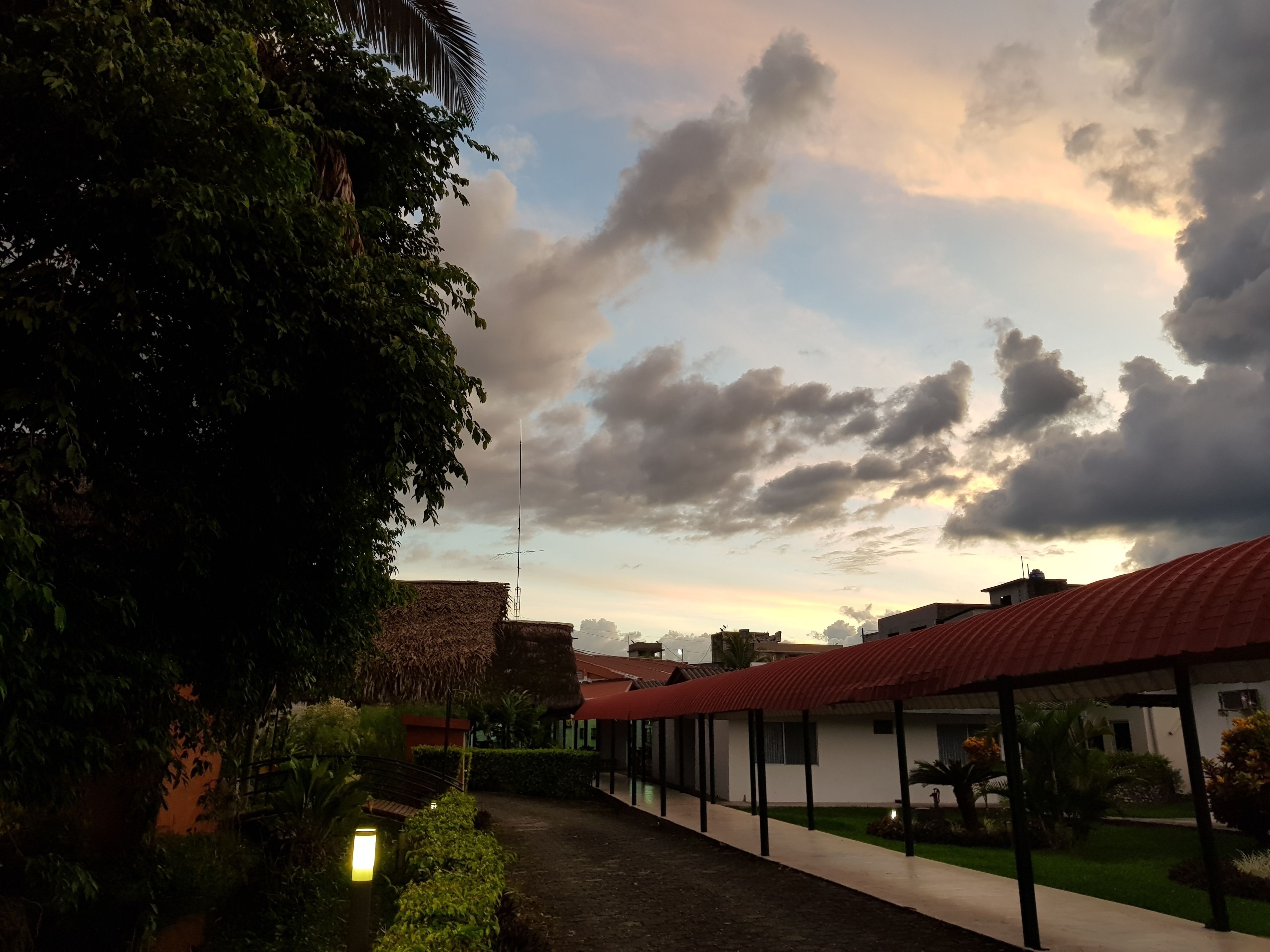 A pathway leading to a building with a red roof, surrounded by greenery and a clear sky with clouds.