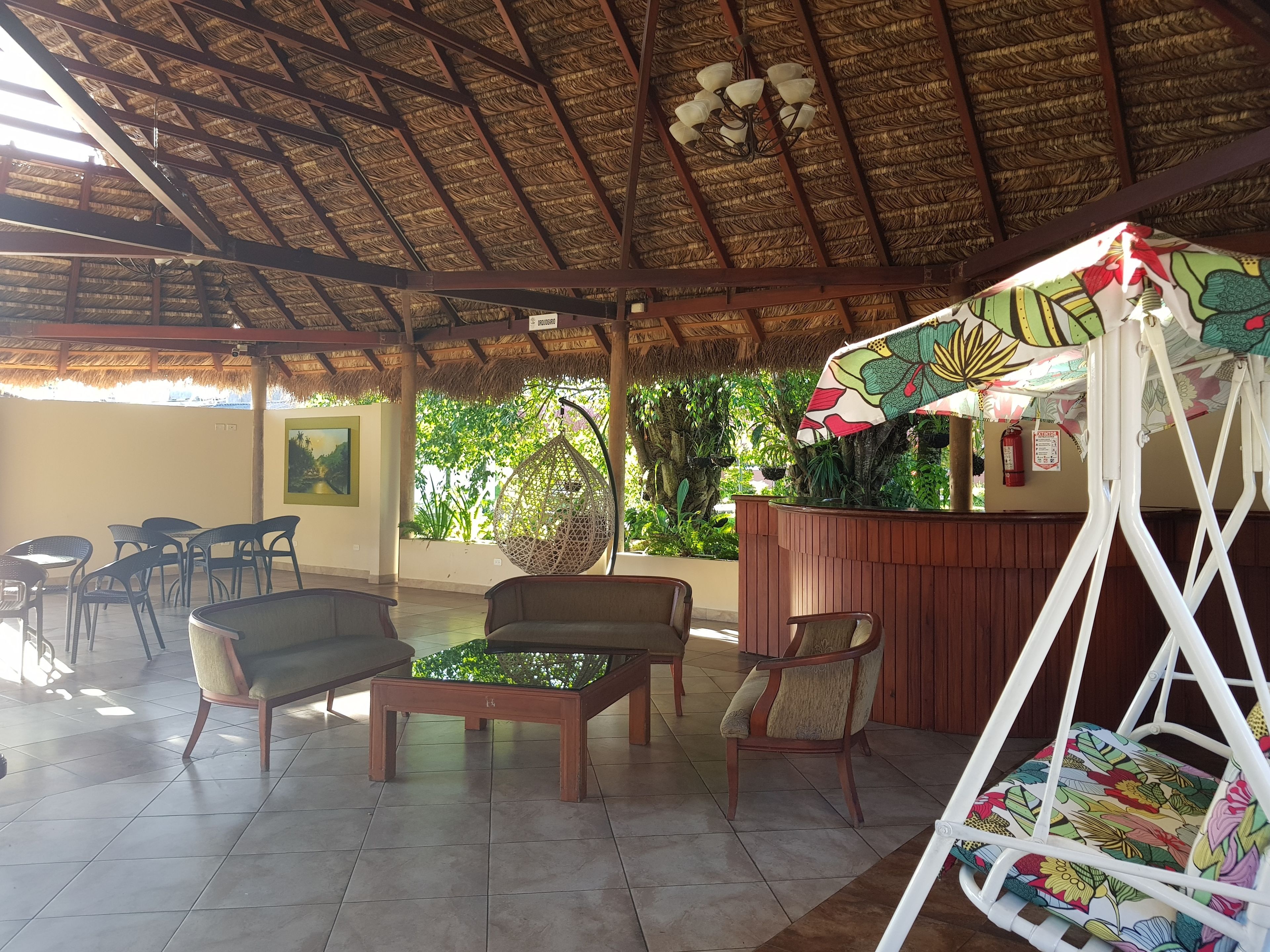 Un espace extérieur abrité, aménagé avec des chaises et des tables, un fauteuil suspendu et un parasol coloré.