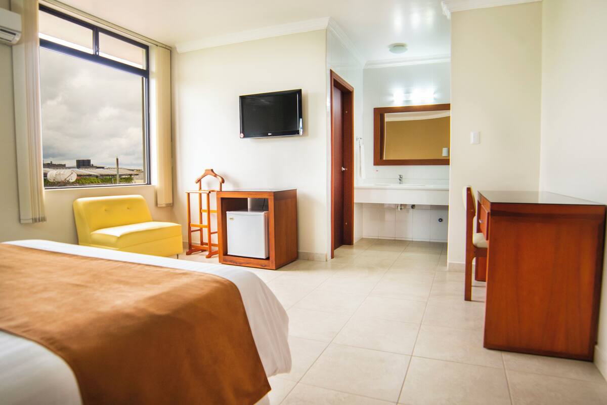 Een hotelkamer met een bed, bureau, stoel, televisie, badkamer met wastafel en spiegel, en een raam met uitzicht op gebouwen.