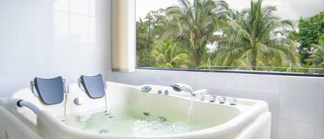 Baño con una bañera grande, dos reposapiés azules y una ventana con vista a palmeras.