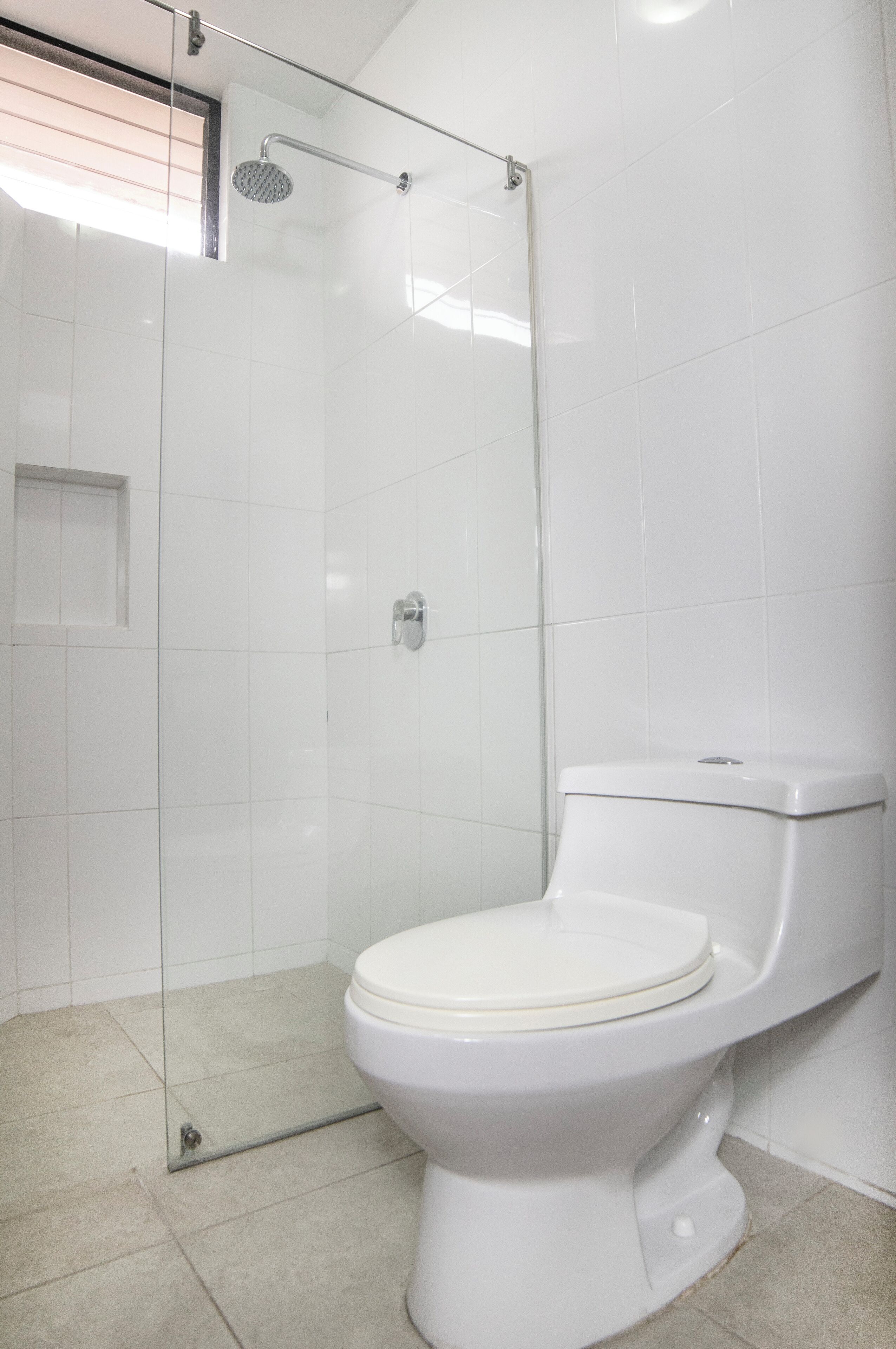 Un baño con inodoro blanco, ducha con mampara de vidrio y paredes con azulejos blancos.