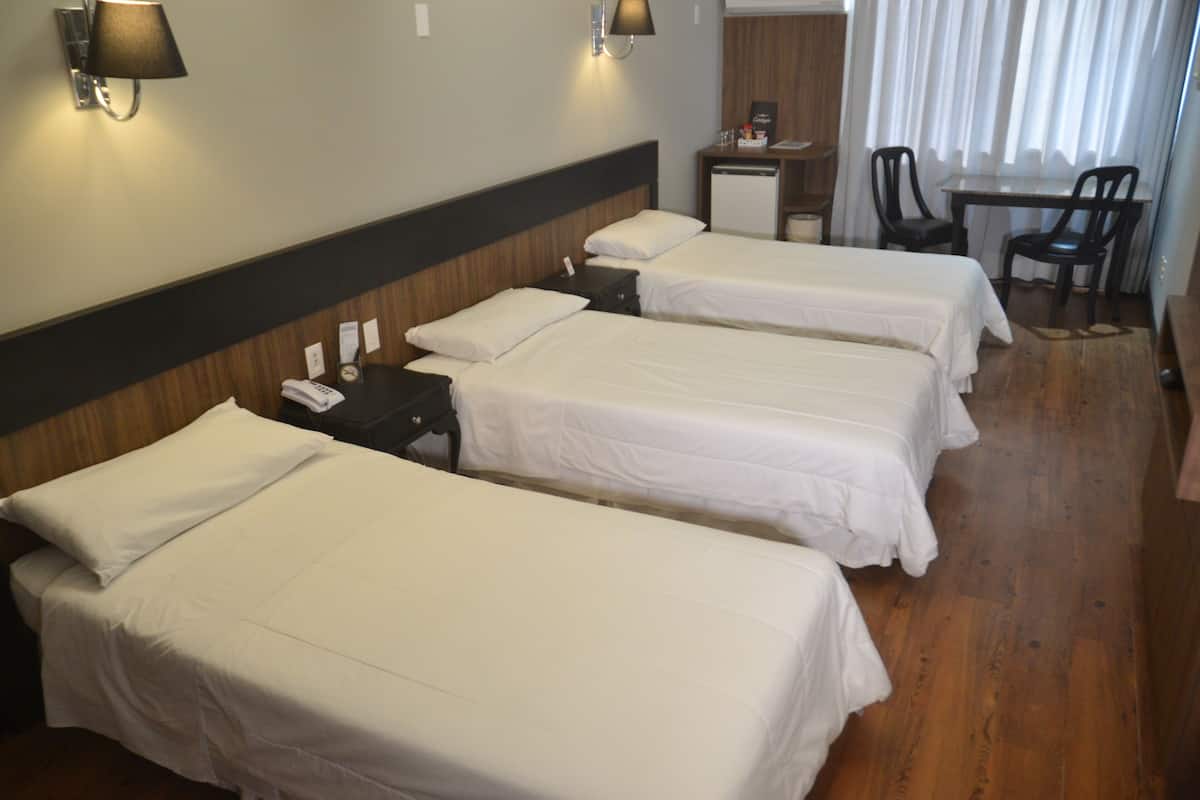 Ein Hotelzimmer mit drei Einzelbetten, einem Schreibtisch mit Stuhl, einer Lampe und einem kleinen Tisch ebenfalls mit einer Lampe.