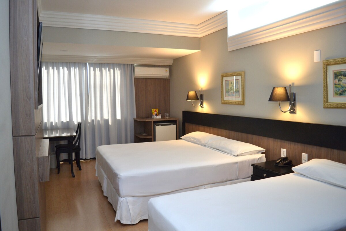Une chambre d’hôtel avec deux lits, un bureau, une chaise, une petite table et une table de chevet avec une lampe.