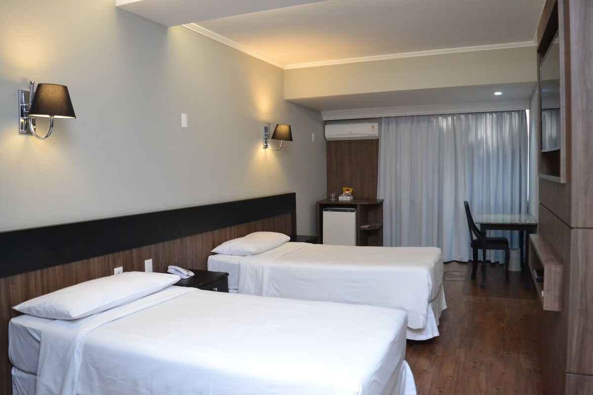 Une chambre d’hôtel avec deux lits, un bureau, une chaise, un miroir et une fenêtre avec des rideaux.