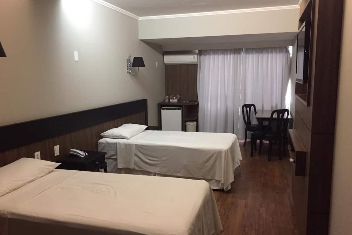 A hotel room with two single beds, a desk with a chair, a small table, a lamp, and a TV.