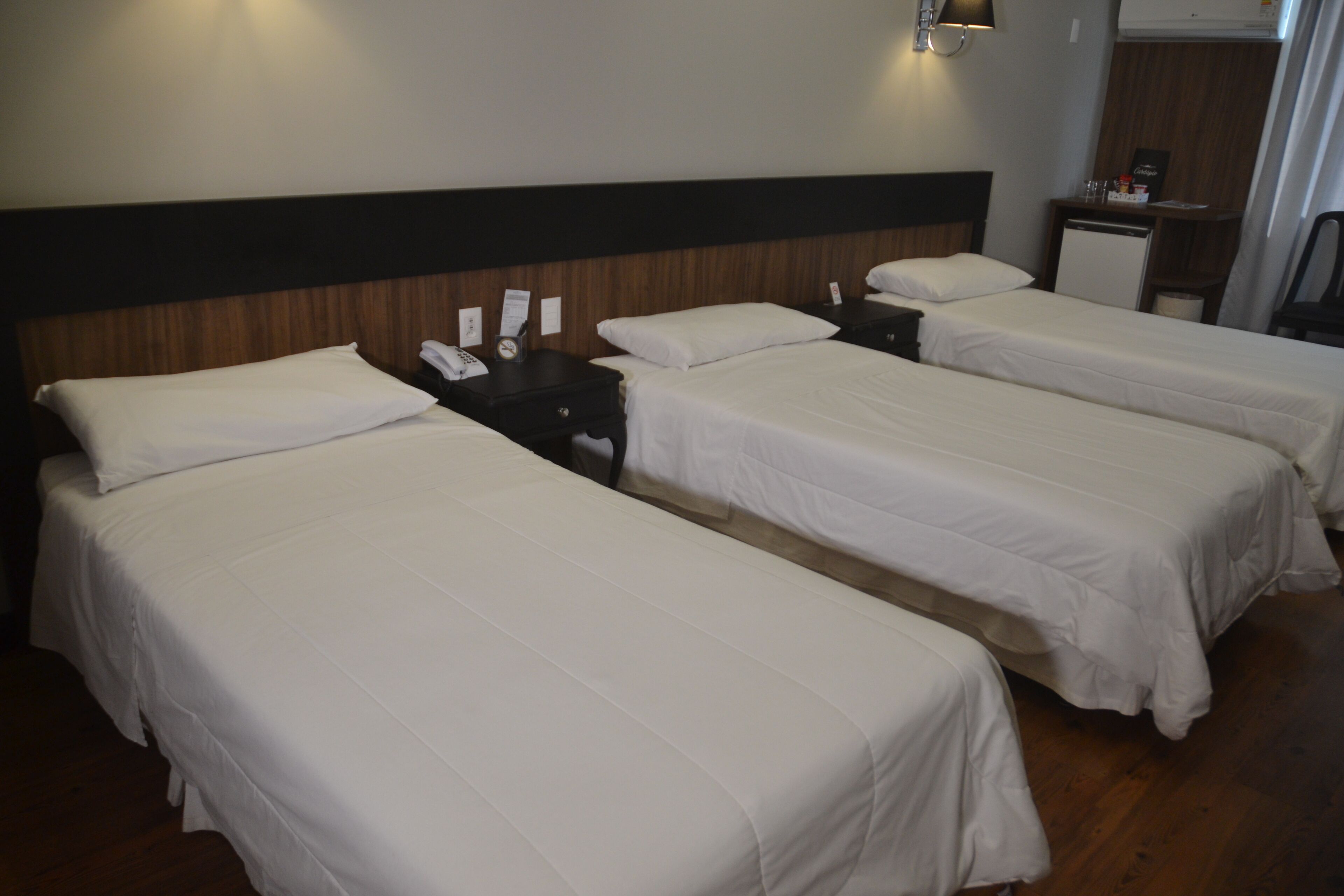 Habitación de hotel con tres camas individuales, cabecera de madera, un escritorio pequeño con teléfono y una mesita de noche con lámpara.