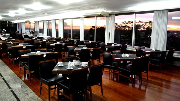 Un restaurante con varias mesas y sillas, ventanas amplias con cortinas y vistas al atardecer.