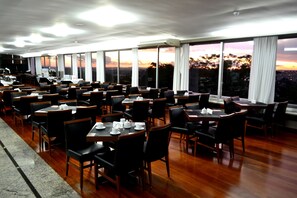 A restaurant with multiple tables and chairs, large windows with curtains, and a view of the sunset. - Premium Vila Velha Ponta Grossa (Ponta Grossa)