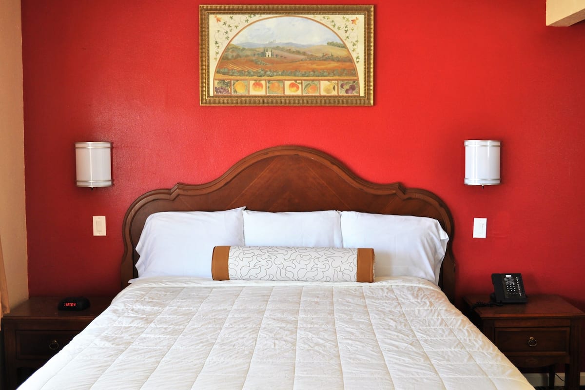 A neatly made bed with a white quilt, a wooden headboard, and a painting above it. There are two bedside tables with lamps and a phone on one of them.