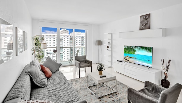 A modern living room with a large TV, a sofa, a coffee table, and a view of the city through large windows. - Ocean Reserve Condominium (Sunny Isles Beach)