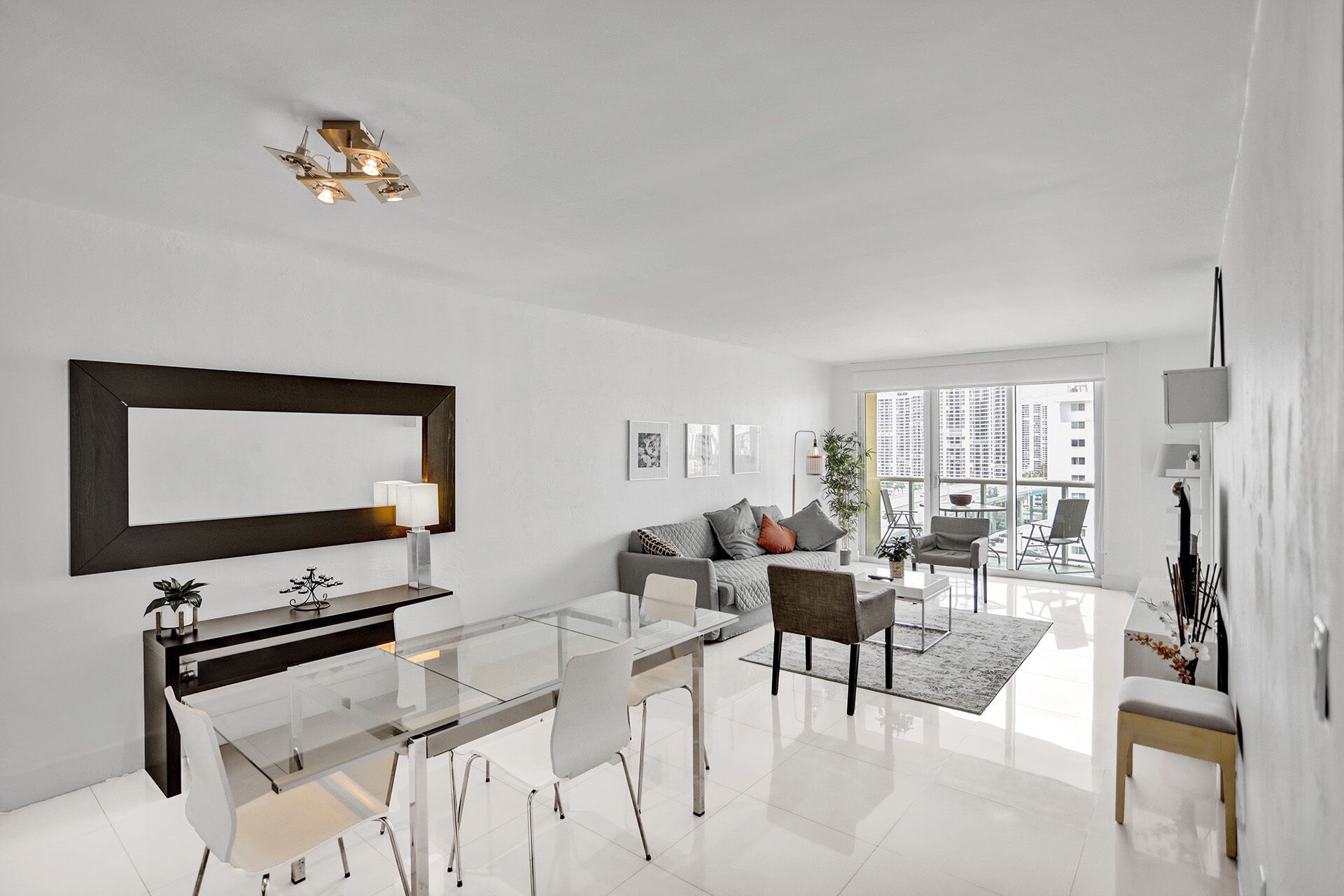 A modern living room with a glass dining table, a sofa, and a coffee table. There is a large mirror on the wall and a ceiling light fixture.