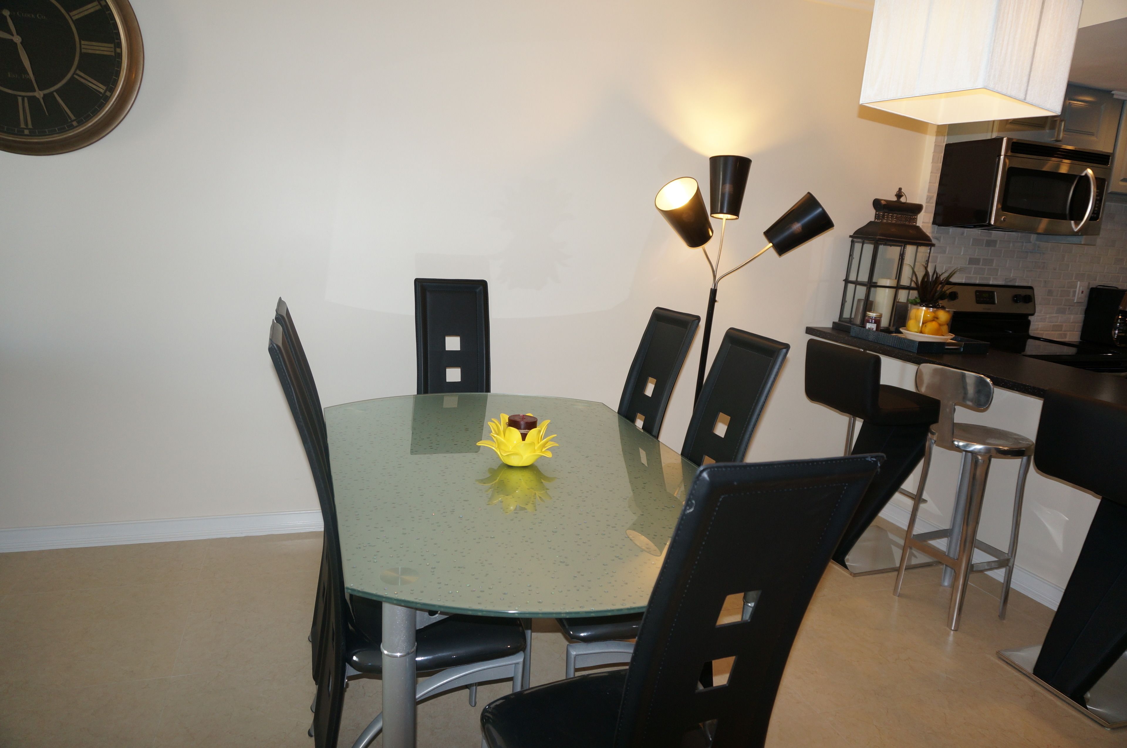 A dining area with a round glass table, black chairs, a wall clock, and a kitchen area with a microwave and a lamp.