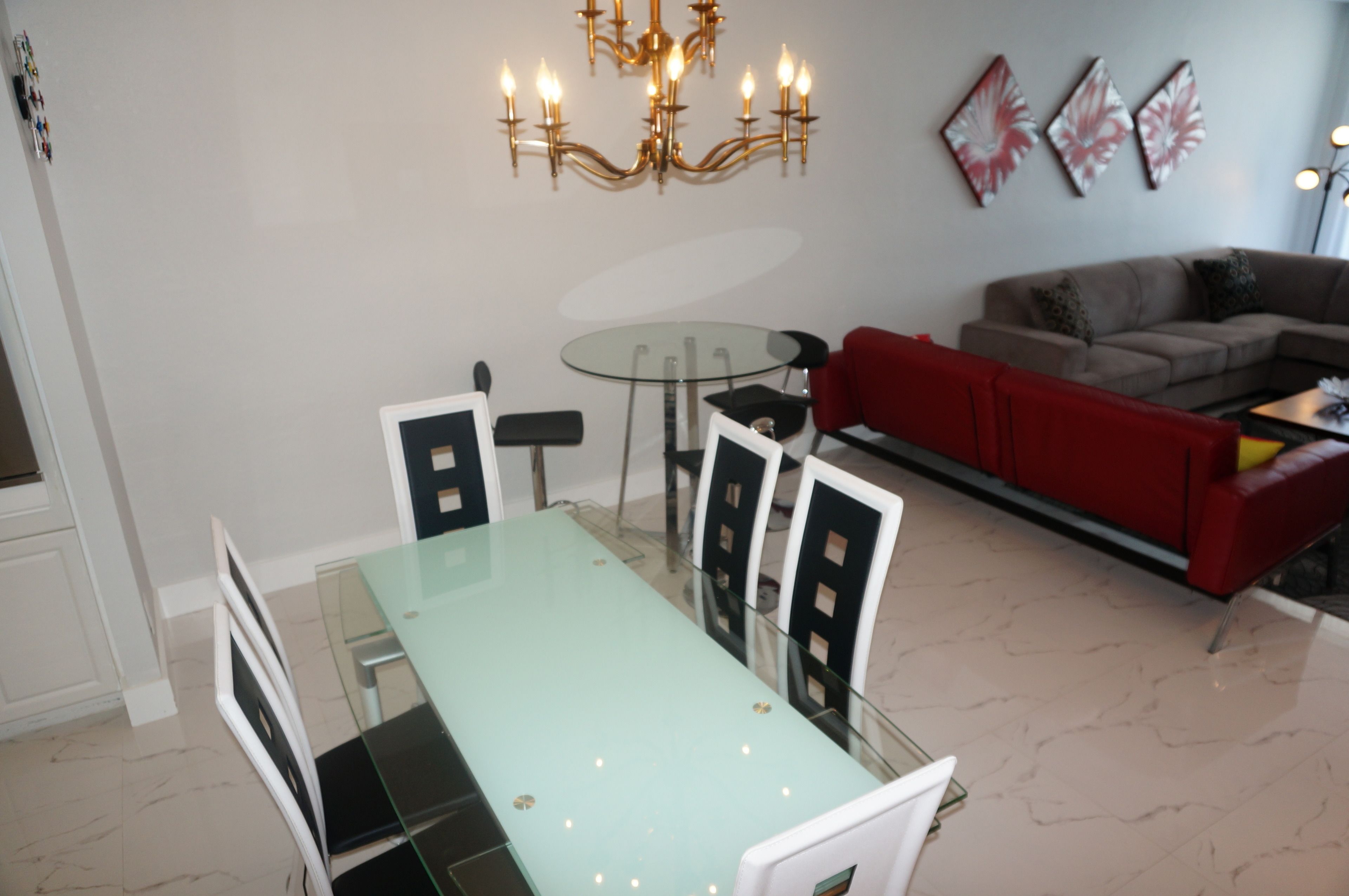 Et spiseområde med et bord med glasplade og stole, en rød sofa og en lysekrone.