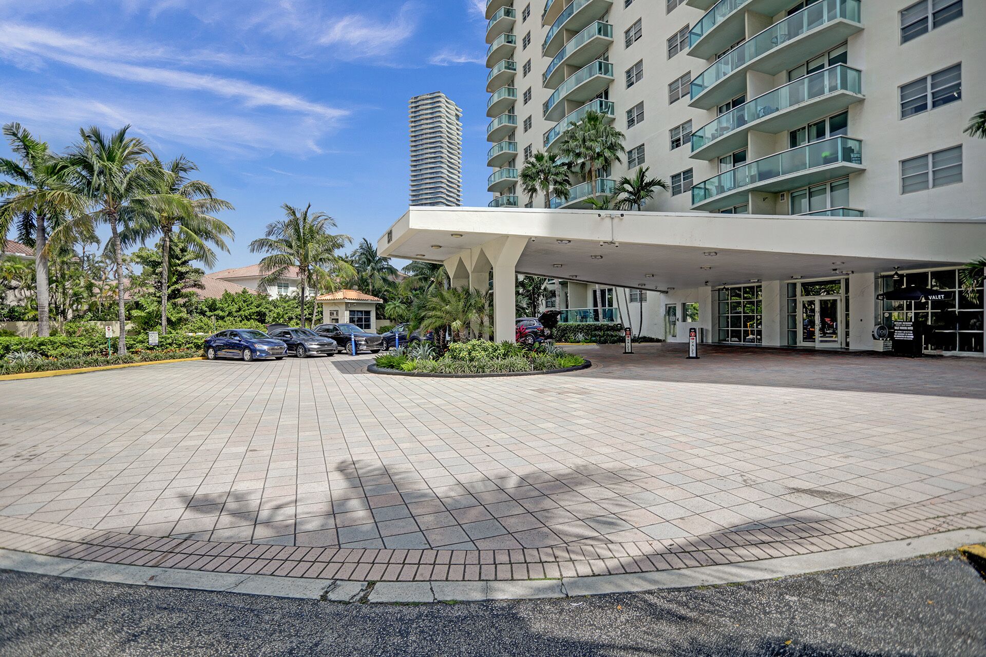 A hotel entrance with a covered walkway, palm trees, and a parking area with cars.