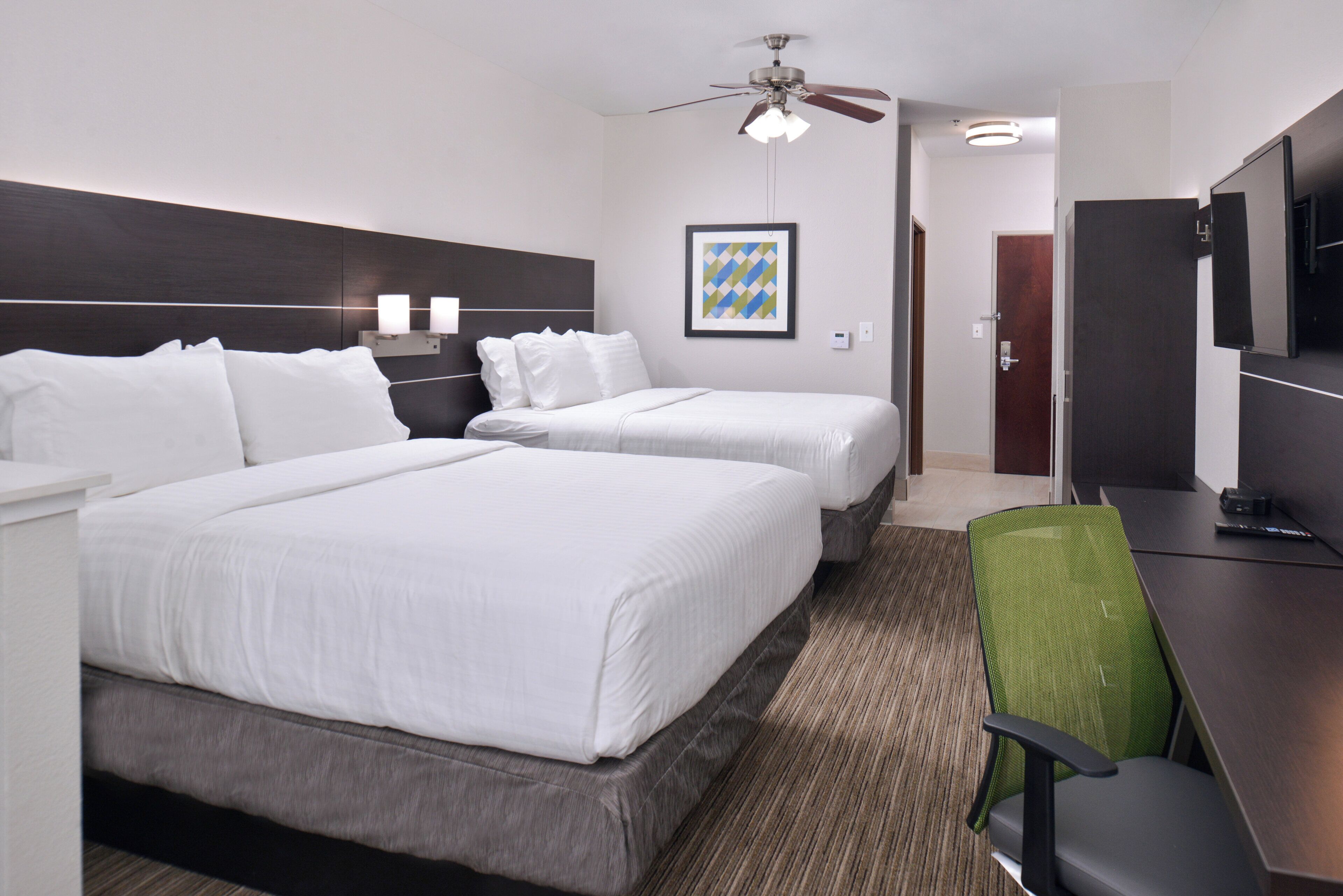 A hotel room with two beds, a desk, a chair, a ceiling fan, and a framed picture on the wall.