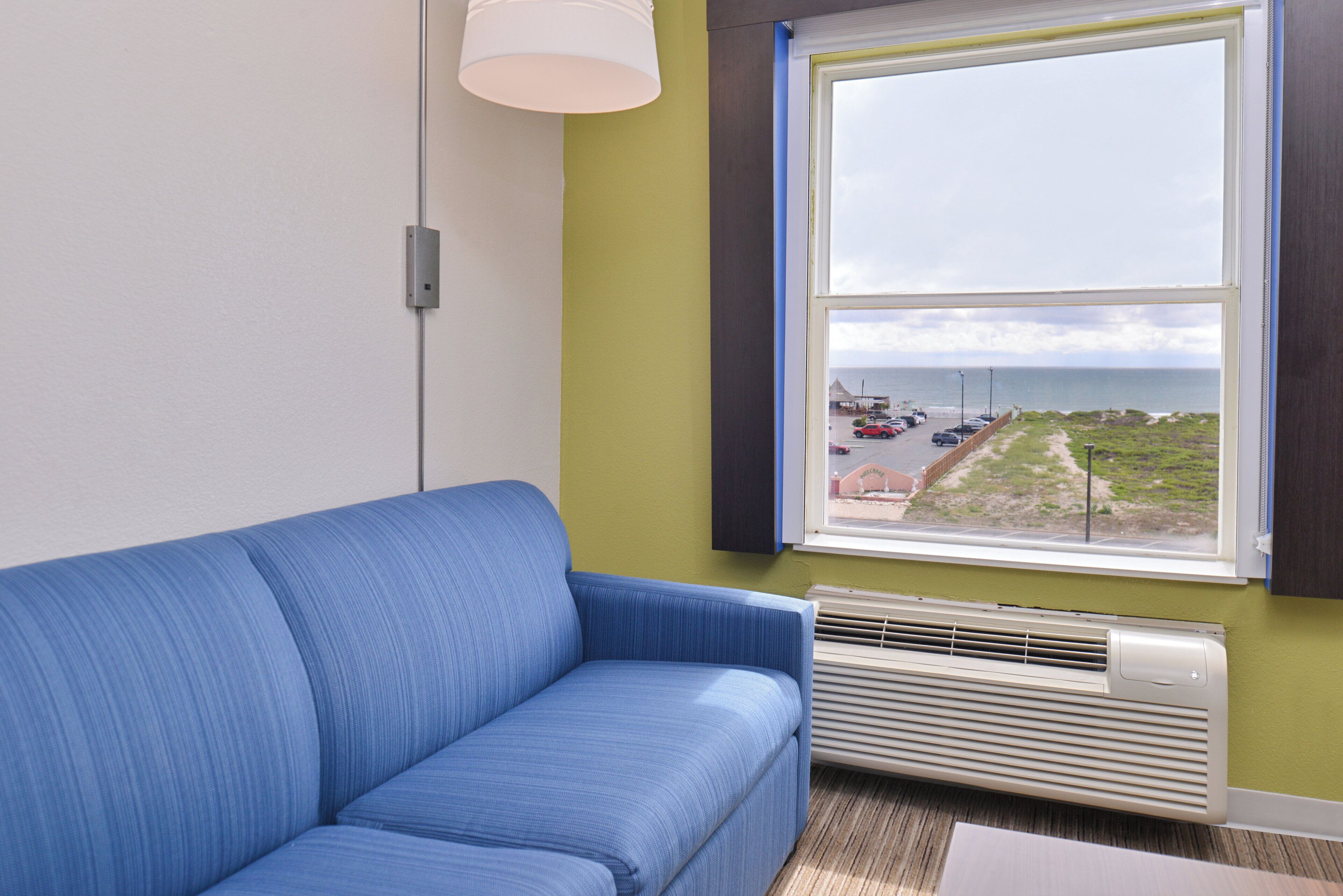 A room with a blue sofa, a window with a view of a beach and parked cars, and a wall-mounted air conditioner.