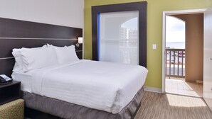 Une chambre d’hôtel comprenant un lit, une table de chevet, un pouf à motifs, une porte donnant sur un balcon, et une vue sur des immeubles et un parking.