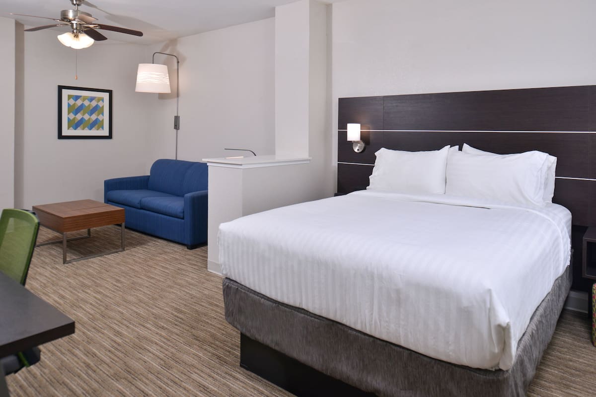 Ein Hotelzimmer mit einem großen Bett, einem blauen Sessel, einem kleinen Tisch, einem Deckenventilator und einer Wandlampe.