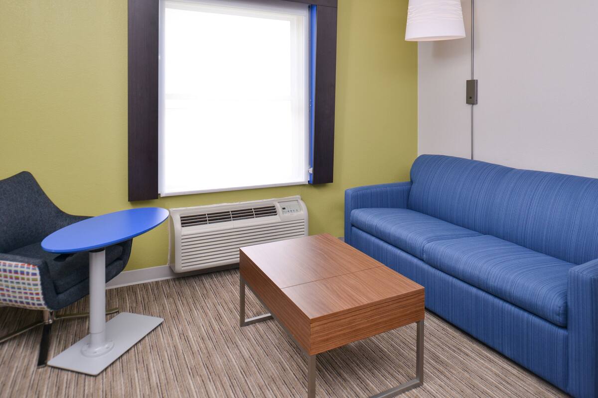Ein Zimmer mit blauem Sofa, einem kleinen runden Tisch, einem Holztisch und einem Fenster mit Jalousien sowie einer Stehlampe.