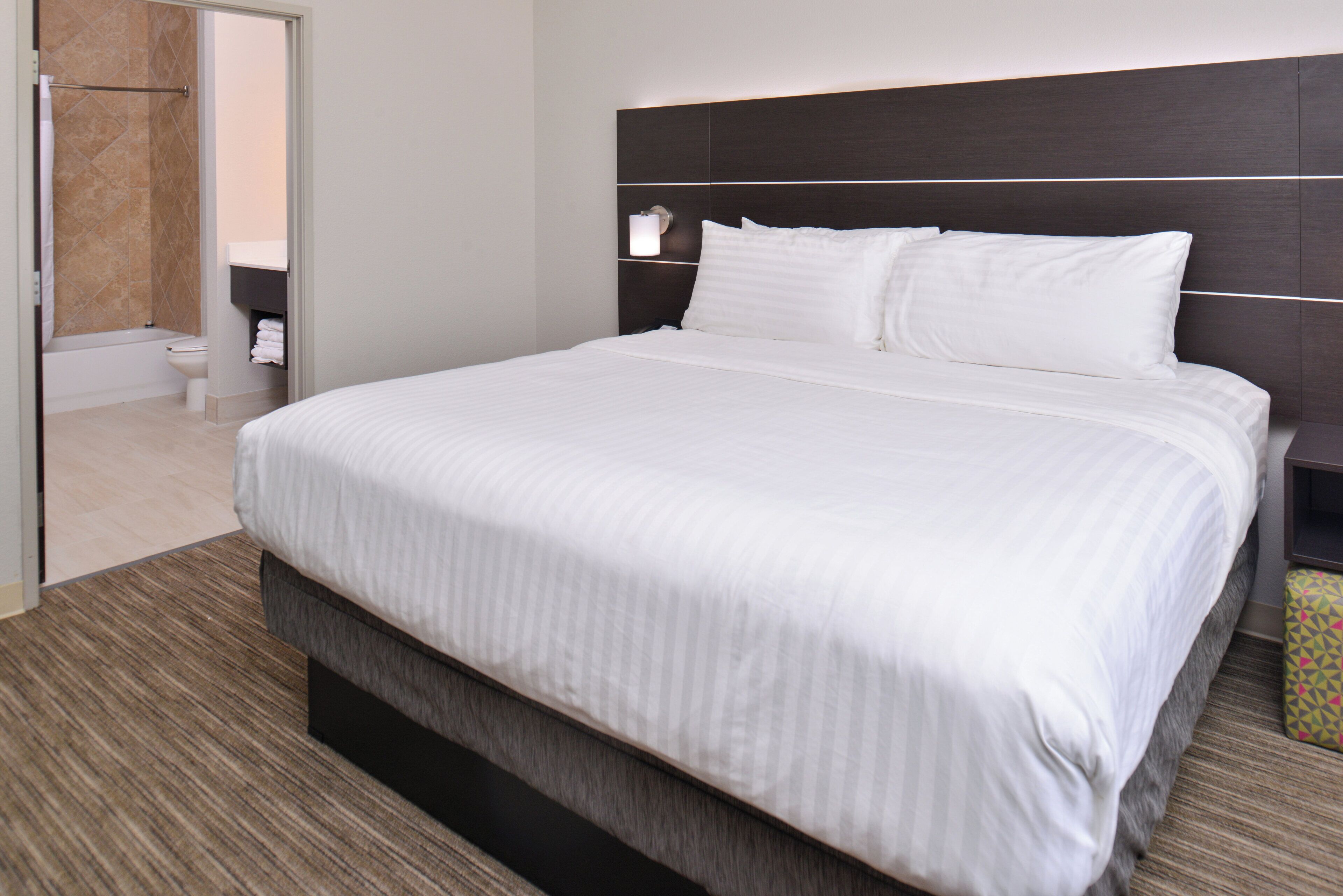 Habitación de hotel con una cama grande, cabecera, mesita de noche y baño visible a través de una puerta abierta.