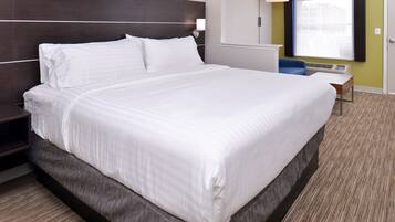A large bed with white linens, a dark headboard, a bedside table, a lamp, and a window with blinds.
