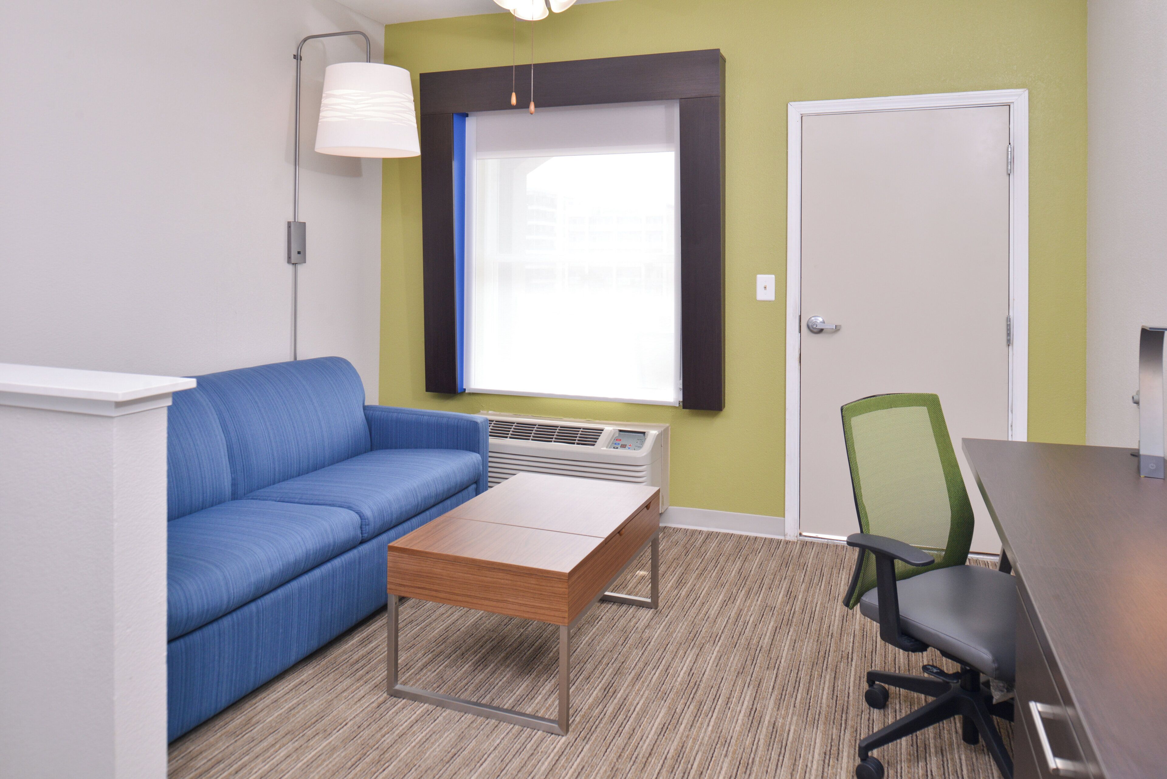 Una habitación con un sofá azul, una mesa de madera, una silla de oficina verde con red, una puerta blanca y una ventana con persianas.