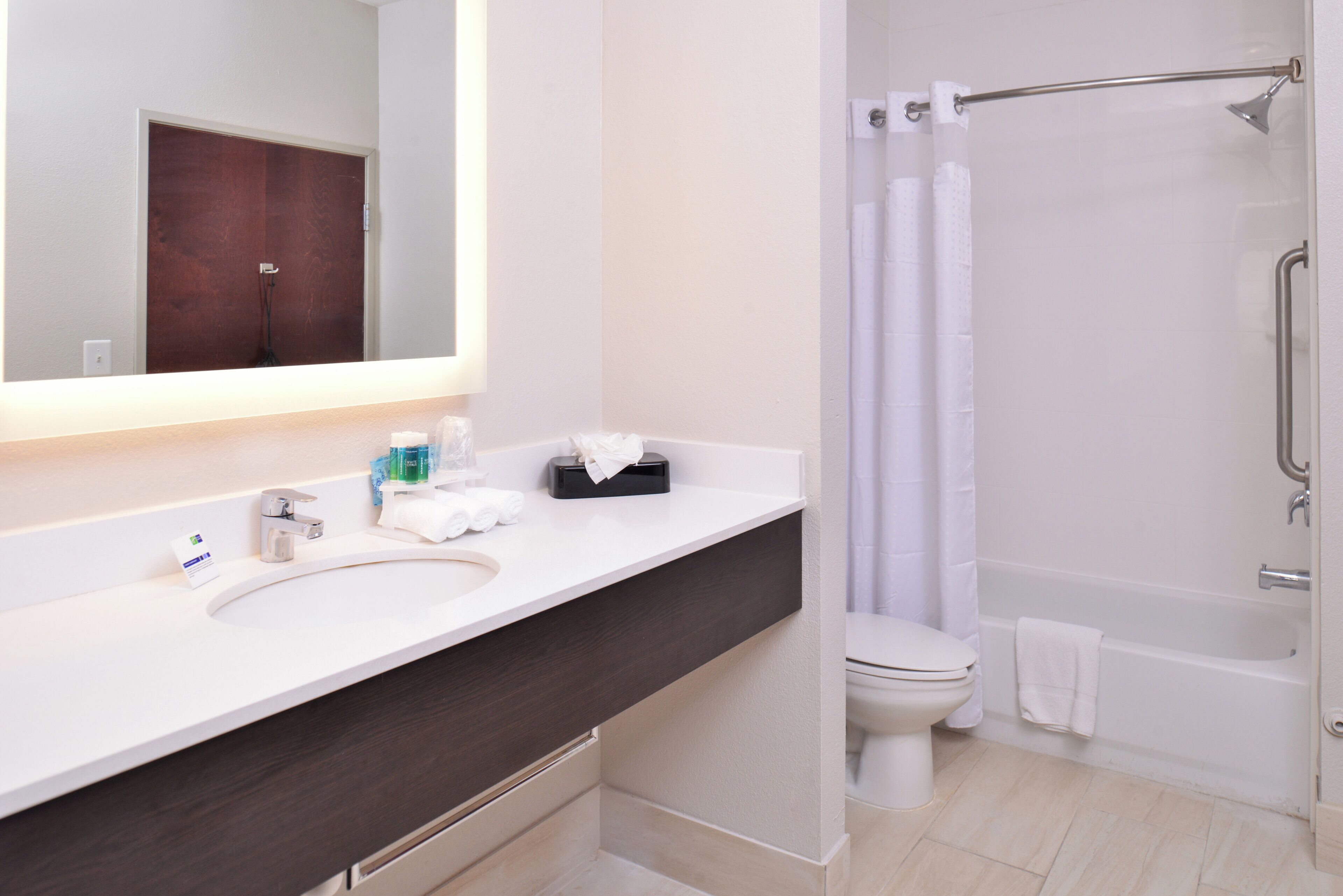 Un baño moderno con lavamanos blanco, tocador de madera oscura, un espejo, inodoro y ducha con puerta de vidrio.