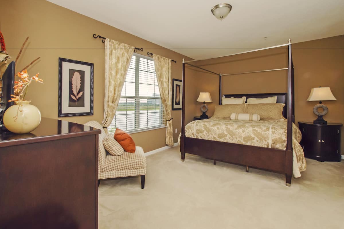 A four-poster bed with a floral bedspread, a desk with a vase, and a chair with a cushion.