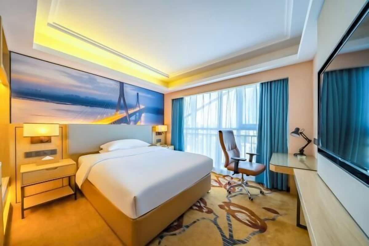 A hotel room with a bed, a desk, a chair, a TV, and a painting on the wall.